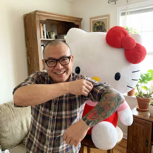 Middle-Aged Man with Glasses and Tattoo Standing by Cartoon Cat Plush Toy